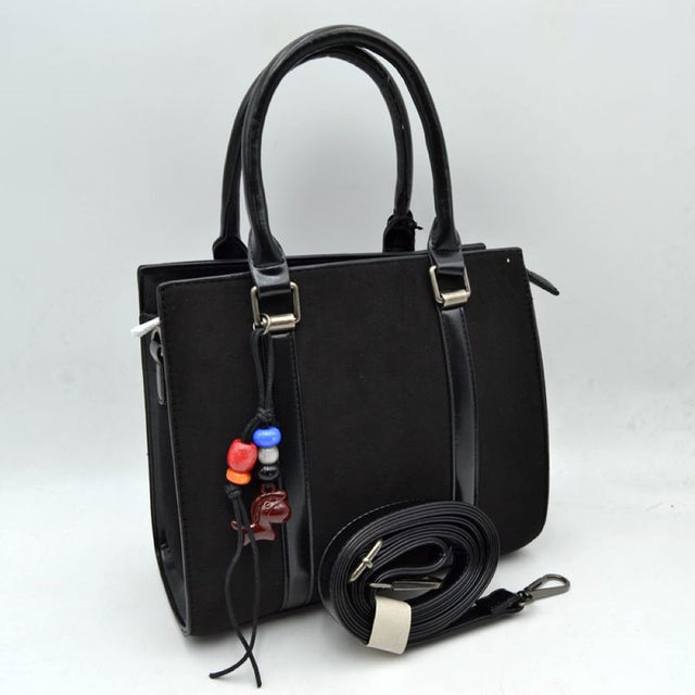 Black This sophisticated satchel pairs a smooth matte main panel with sleek, polished faux leather trim for a classic mixed-media look. It features a structured, rectangular silhouette equipped with sturdy dual top-roll handles anchored by metallic square loops. A decorative knotted cord charm featuring colorful accent beads and a stylized animal pendant hangs from the front handle base, adding a subtle playful note. Designed for versatile styling, the bag includes a main zippered top closure and comes comp